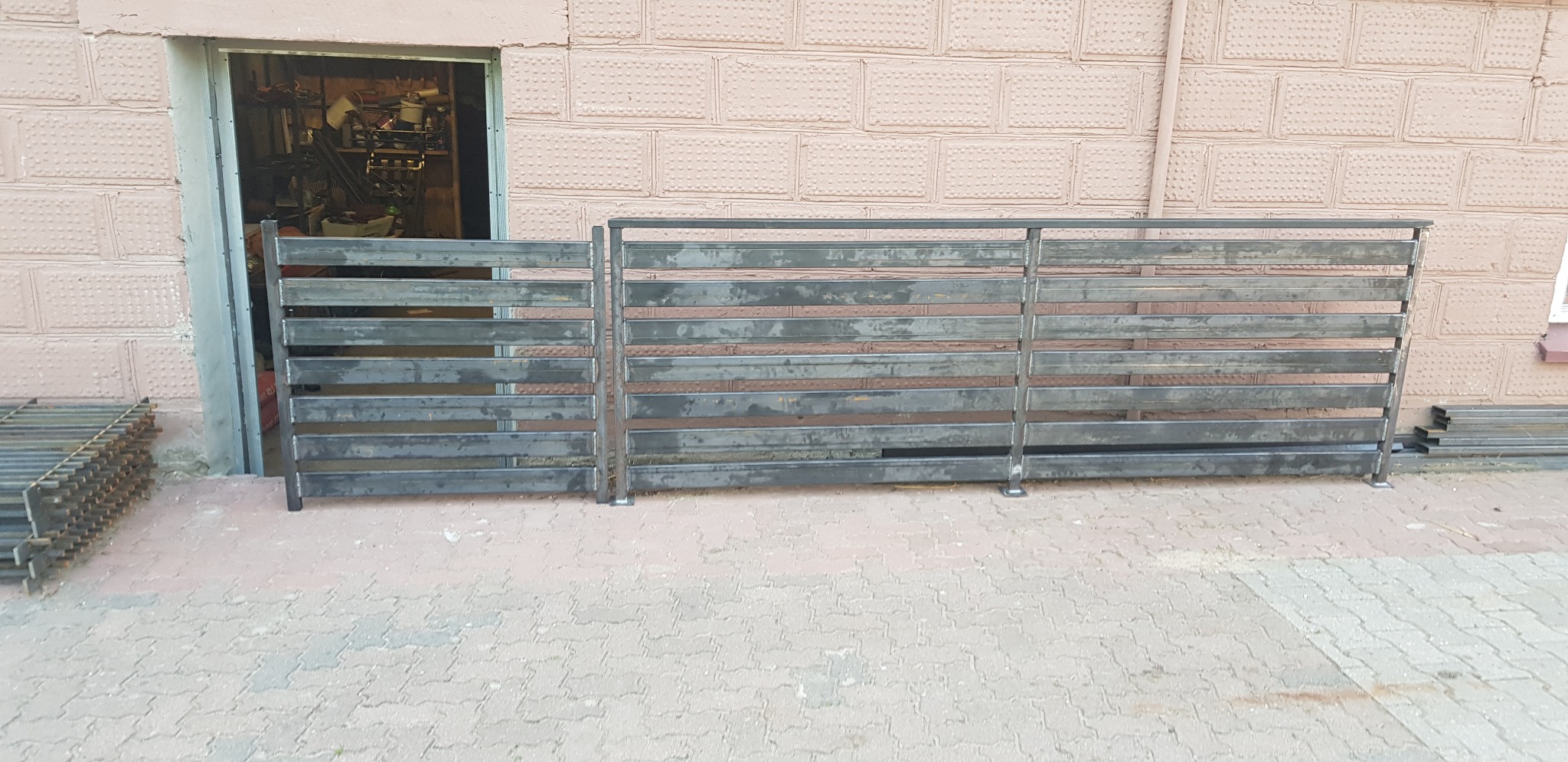 Surowe, niepomalowane przęsła ogrodzenia metalowego z poziomych profili, przygotowane do montażu, składowane przed budynkiem z cegły.