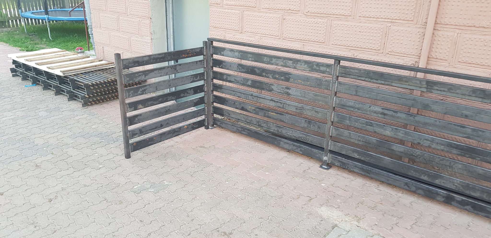 Surowe, metalowe segmenty ogrodzenia z poziomymi szczeblami, przygotowane do montażu, ułożone na kostce brukowej obok ściany budynku.