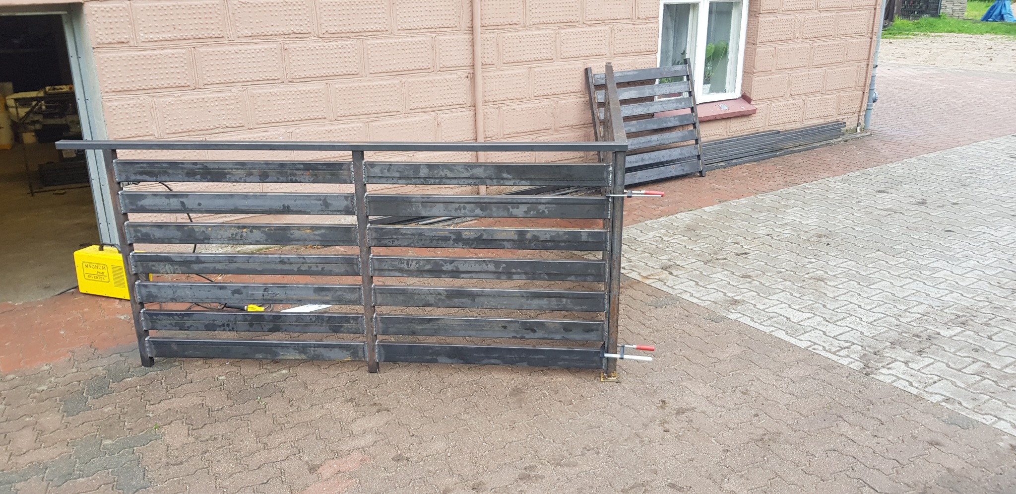Surowa, spawana konstrukcja metalowa ogrodzenia z poziomymi szczeblami, częściowo zamocowana zaciskami, obok leży spawarka transformatorowa, w tle budynek z cegły i okno.