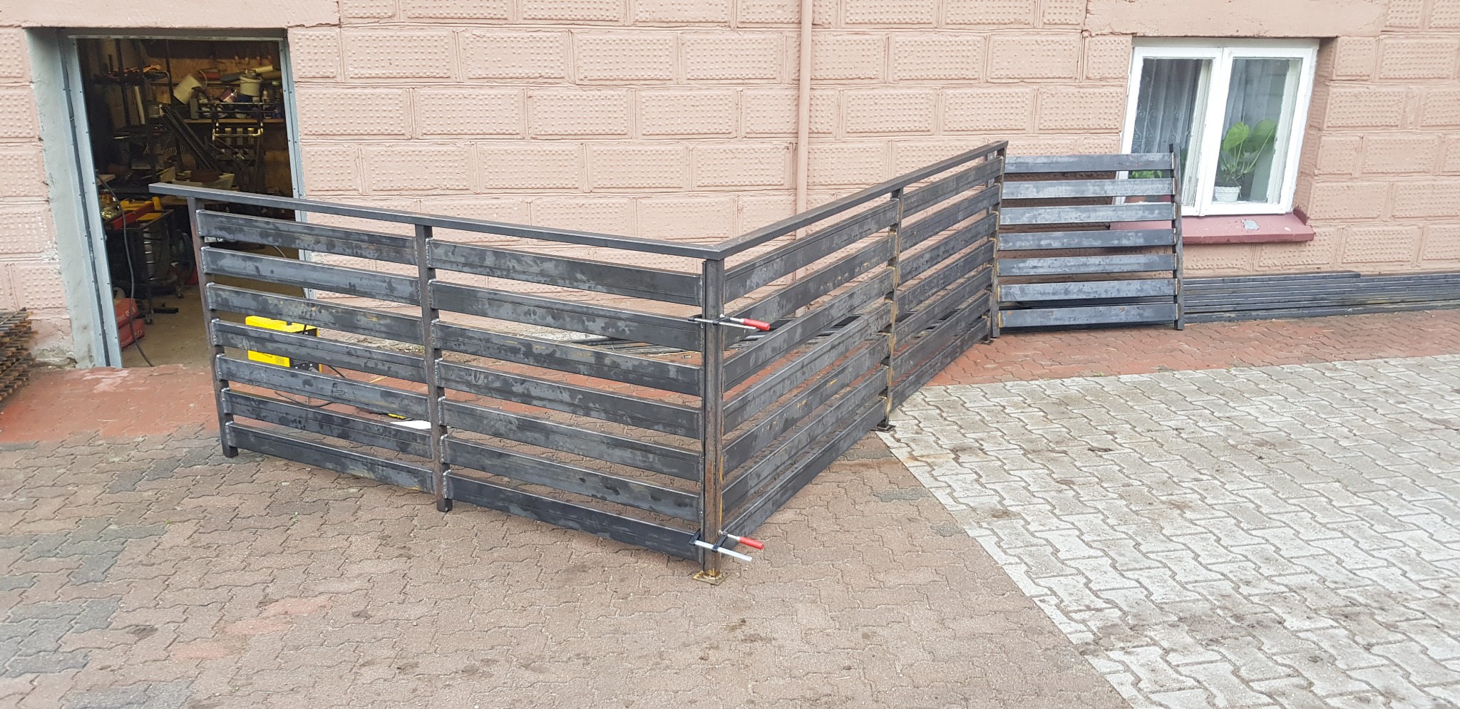 Surowy, metalowy szkielet ogrodzenia z poziomych profili, częściowo zmontowany na zewnątrz budynku z cegły, z widocznymi ściskami stolarskimi i żółtą spawarką w tle warsztatu.