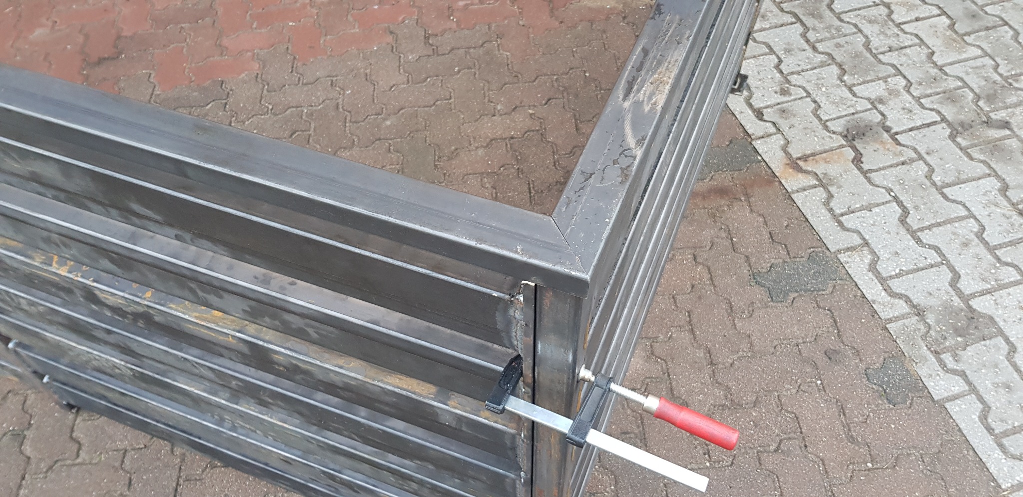 Fragment konstrukcji metalowej z profili zamkniętych, spawana, z widocznym śladem spawania i zaciskiem śrubowym z czerwoną rączką, prawdopodobnie w trakcie montażu.