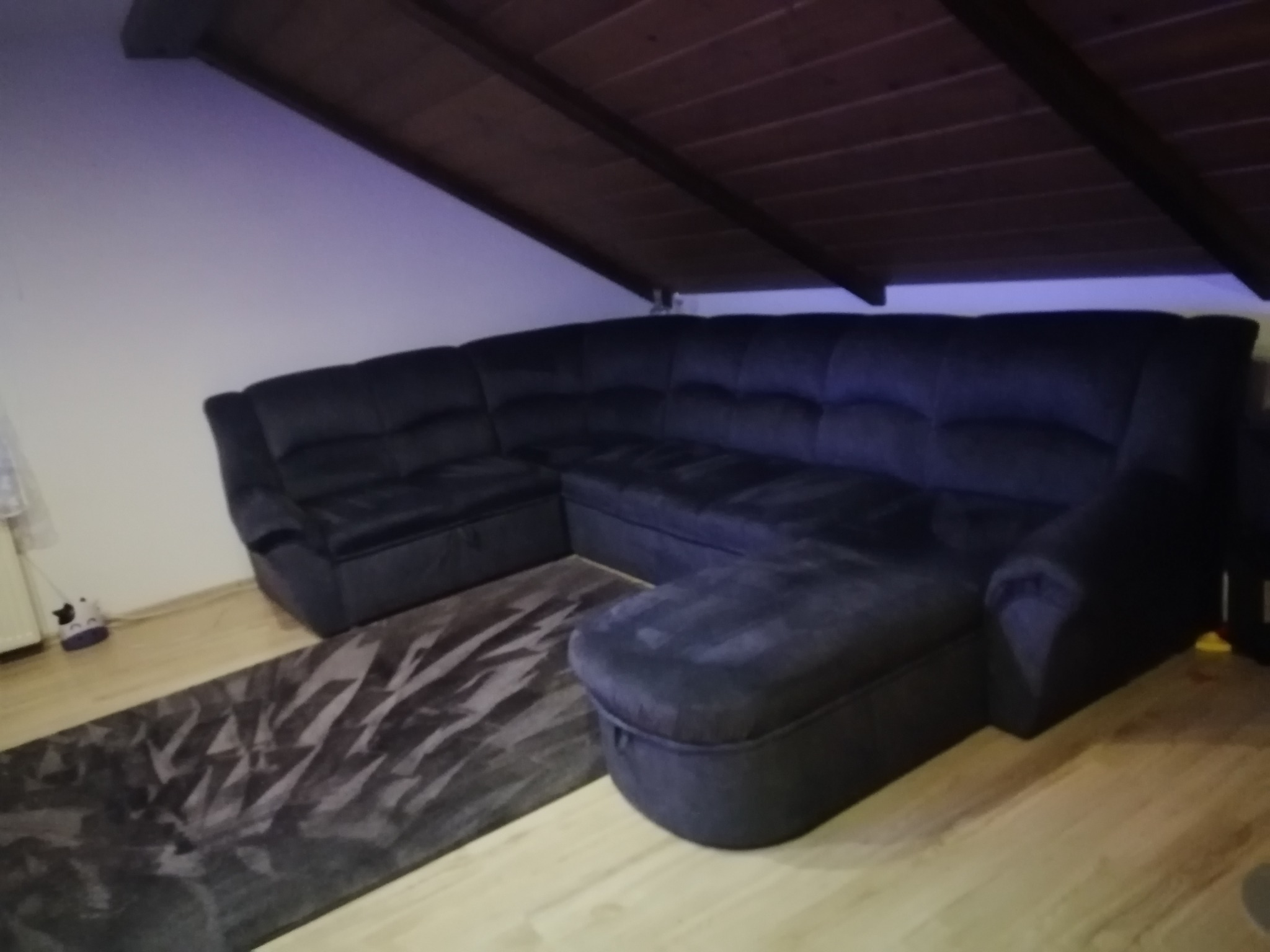 Ciemnoszara, welurowa sofa narożna z pufą na tle ukośnego sufitu i wzorzystego dywanu, w pomieszczeniu oświetlonym fioletowym światłem.