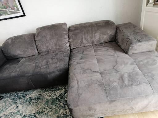 Szara, modułowa kanapa narożna z widocznymi plamami po czyszczeniu na jasnej części, kontrastująca z ciemniejszą, nieczyszczoną sekcją po lewej stronie.