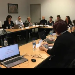 Zespół osób w sali konferencyjnej, dyskusja nad planem komunikacji widocznym na ekranie laptopa, butelki wody na stole, formalna atmosfera.