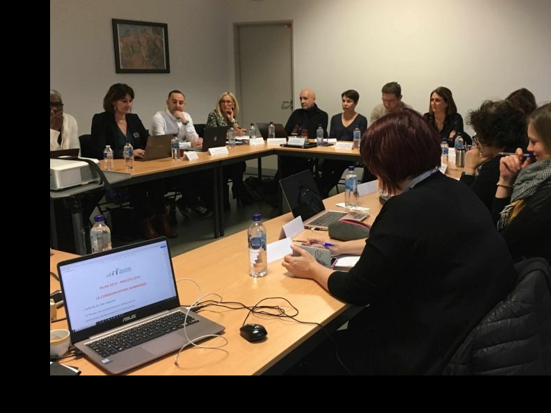 Zespół osób w sali konferencyjnej, dyskusja nad planem komunikacji widocznym na ekranie laptopa, butelki wody na stole, formalna atmosfera.