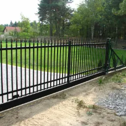 Czarna, metalowa brama przesuwna z automatyką, widoczna z zewnątrz posesji. Brama z profilu kwadratowego, ocynkowana ogniowo i malowana natryskowo, zamontowana na tle zielonego trawnika i drzew.