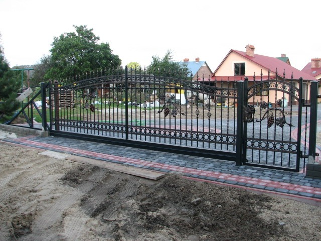 Czarna, metalowa brama przesuwna z motywem roślinnym, ocynkowana i malowana proszkowo, z furtką obok, zamontowana na posesji z kostką brukową i piaskiem.