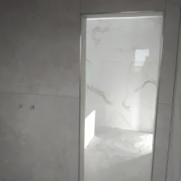 Świeżo wykafelkowana łazienka z szarymi płytkami na ścianach zewnętrznych i białymi marmurkowymi płytkami wewnątrz, widoczne wystające rury instalacji wodnej, podłoga w trakcie kafelkowania.