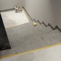 Schody wyłożone szarymi płytkami imitującymi lastryko, zabezpieczone żółtą taśmą malarską, z gąbką na jednym ze stopni i wiadrem z narzędziami w tle.