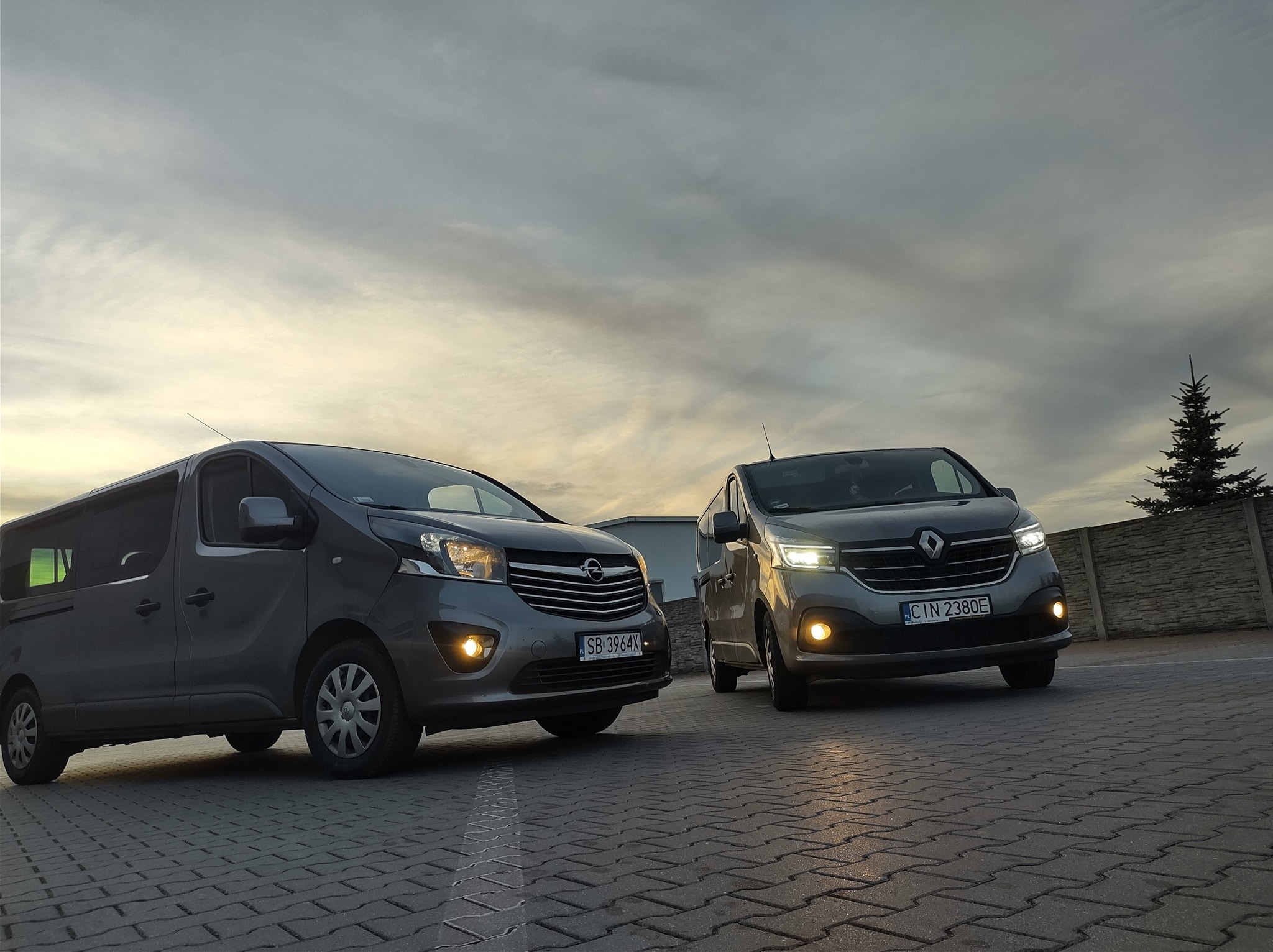 Dwa busy: Opel Vivaro i Renault Trafic, zaparkowane na brukowanej nawierzchni z włączonymi światłami mijania, na tle pochmurnego nieba o zmierzchu.