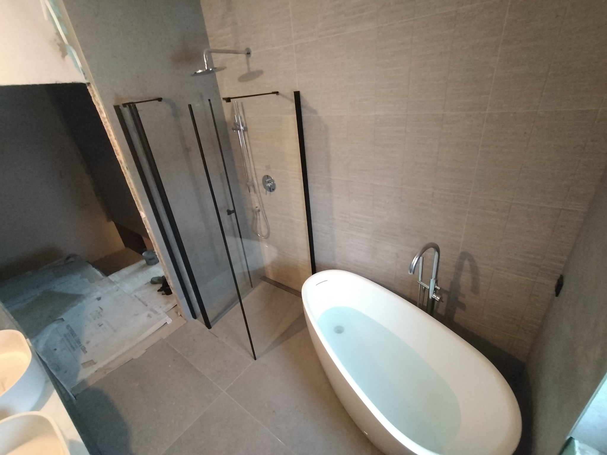 Nowoczesna łazienka w trakcie wykańczania z wolnostojącą wanną wypełnioną wodą i kabiną prysznicową z czarnymi ramami, widoczne płytki na ścianach i podłodze, oraz przygotowane podłoże w strefie...