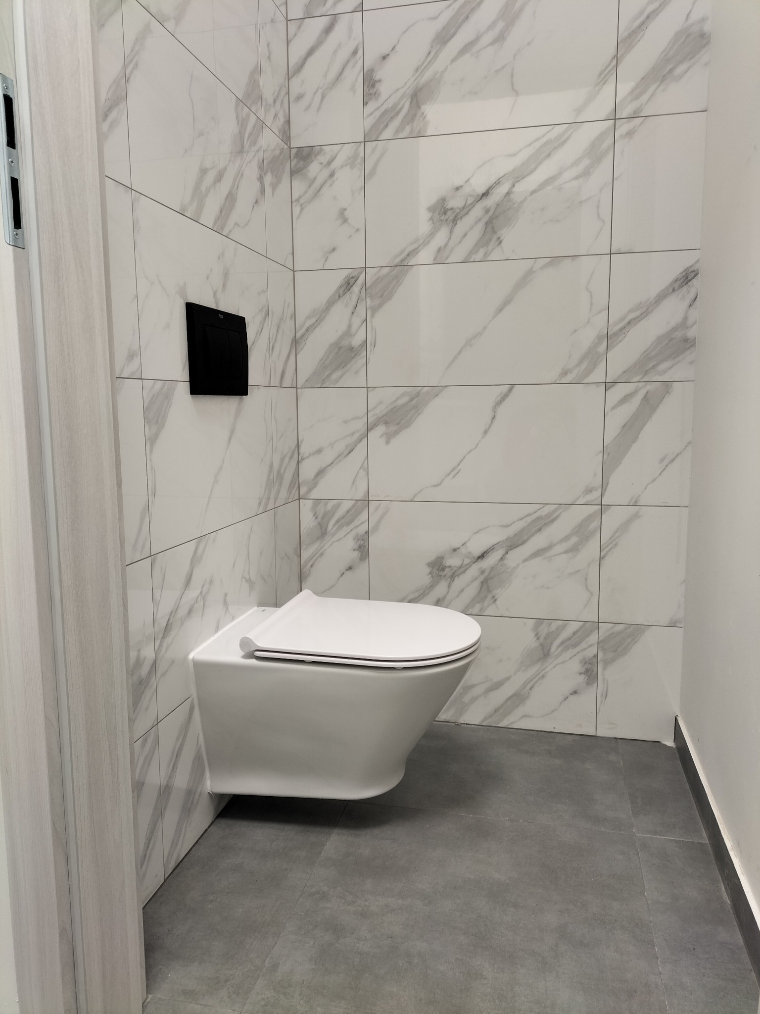 Nowoczesna toaleta podwieszana z czarnym przyciskiem spłukującym na tle marmurowych płytek ściennych i szarej podłogi.