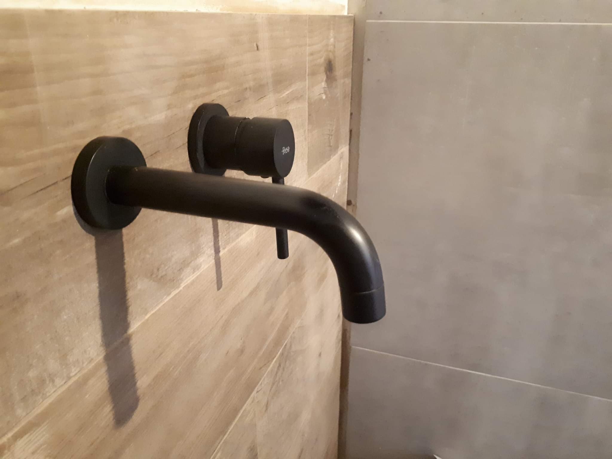 Matowa, czarna bateria umywalkowa marki Rea zamontowana na ścianie wyłożonej drewnopodobnymi płytkami w łazience.