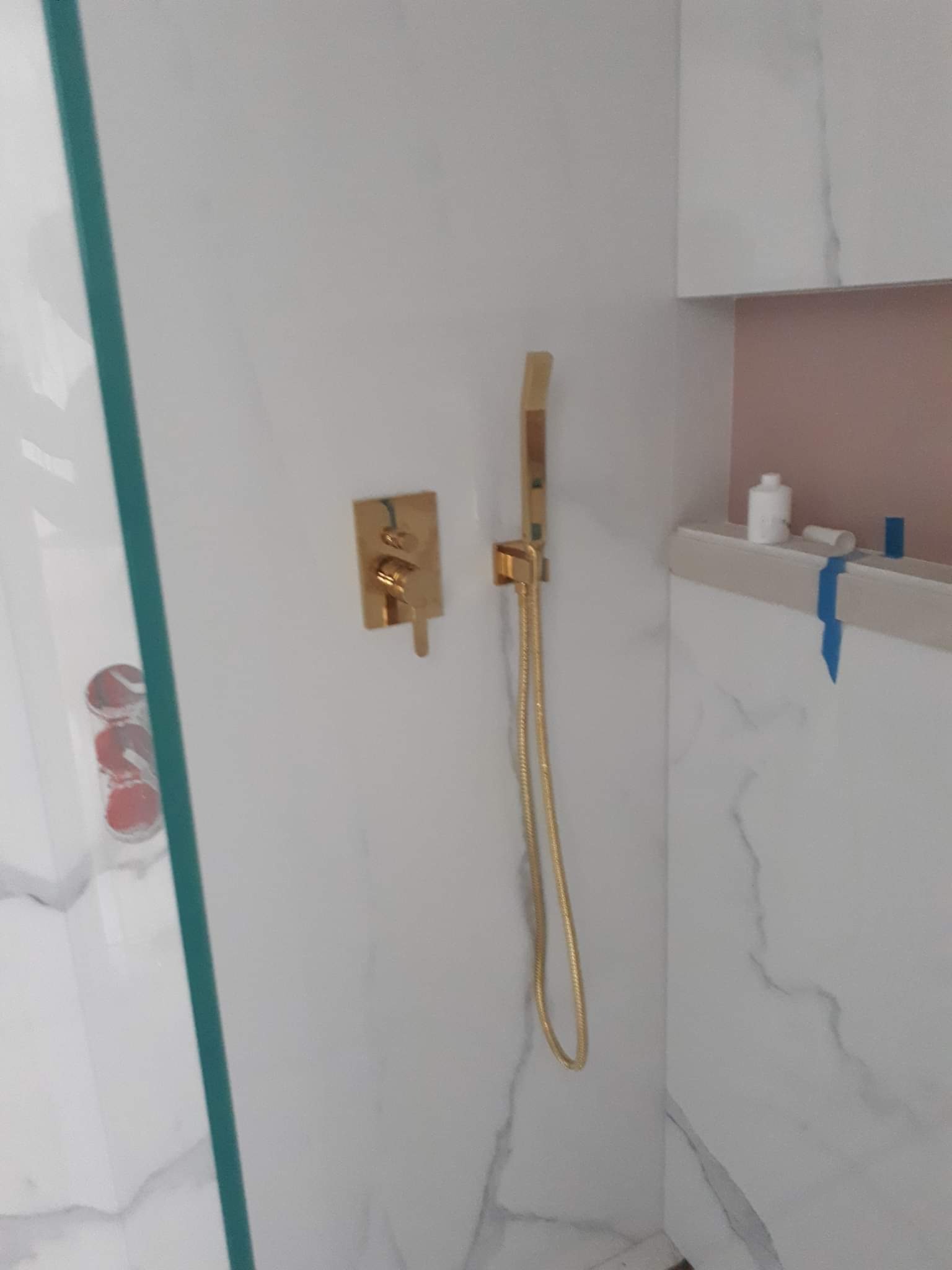 Złota bateria prysznicowa z deszczownicą i termostatem w kabinie wyłożonej białym marmurem z półką na kosmetyki.