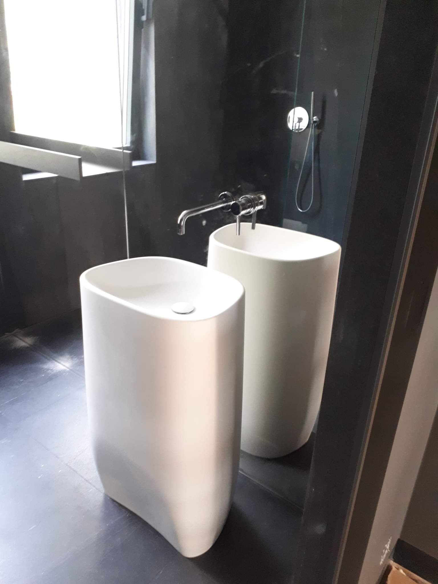 Dwie wolnostojące, owalne umywalki w nowoczesnej łazience z czarnymi ścianami i podłogą, widoczna armatura i kabina prysznicowa.