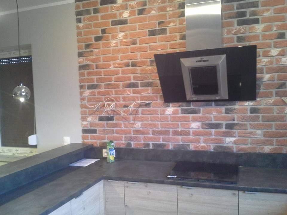 Kuchnia z ceglaną ścianą, czarnym okapem i szarym blatem, widoczne wystające kable, lampa wisząca i butelka na blacie.