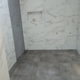 Wykończona łazienka z szarymi płytkami podłogowymi i białymi marmurkowymi płytkami ściennymi, wnęka z dekoracją i odpływ liniowy.