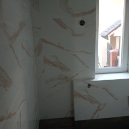 Zakład Remontowo-budowlany Łukasz Rebman - Ściany łazienki wyłożone białymi płytkami z marmurkowym wzorem w odcieniach beżu, widoczne niedokończone wykończenia przy suficie i oknie, czarne wiadro w rogu pomieszczenia.