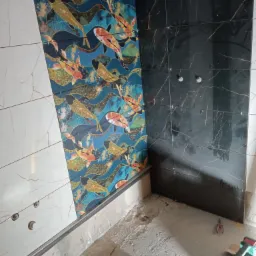 Ściana łazienki z częściowo ułożonymi płytkami: białe marmurkowe, niebieskie z motywem ryb koi i czarne z żyłkowaniem, widoczne narzędzia glazurnicze i niedokończona podłoga.