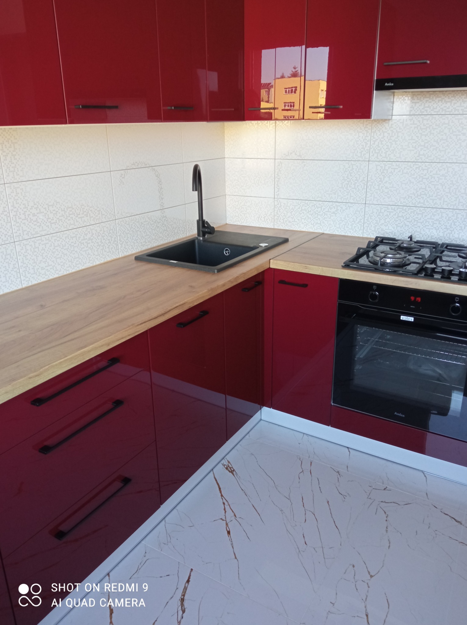 Nowoczesna kuchnia z błyszczącymi, bordowymi szafkami, drewnianym blatem i białymi płytkami mozaikowymi na ścianie, czarny zlew i płyta gazowa, marmurowe płytki podłogowe.