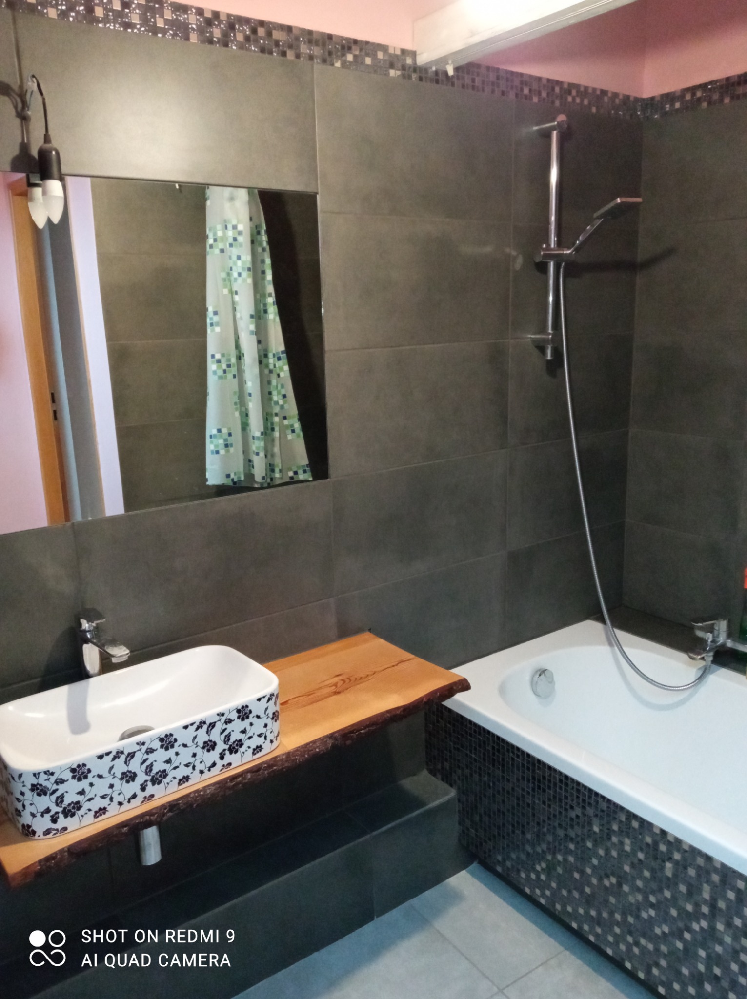 Nowoczesna łazienka z szarymi płytkami, umywalką nablatową w kwiaty na blacie z surowego drewna i wanną obłożoną mozaiką.