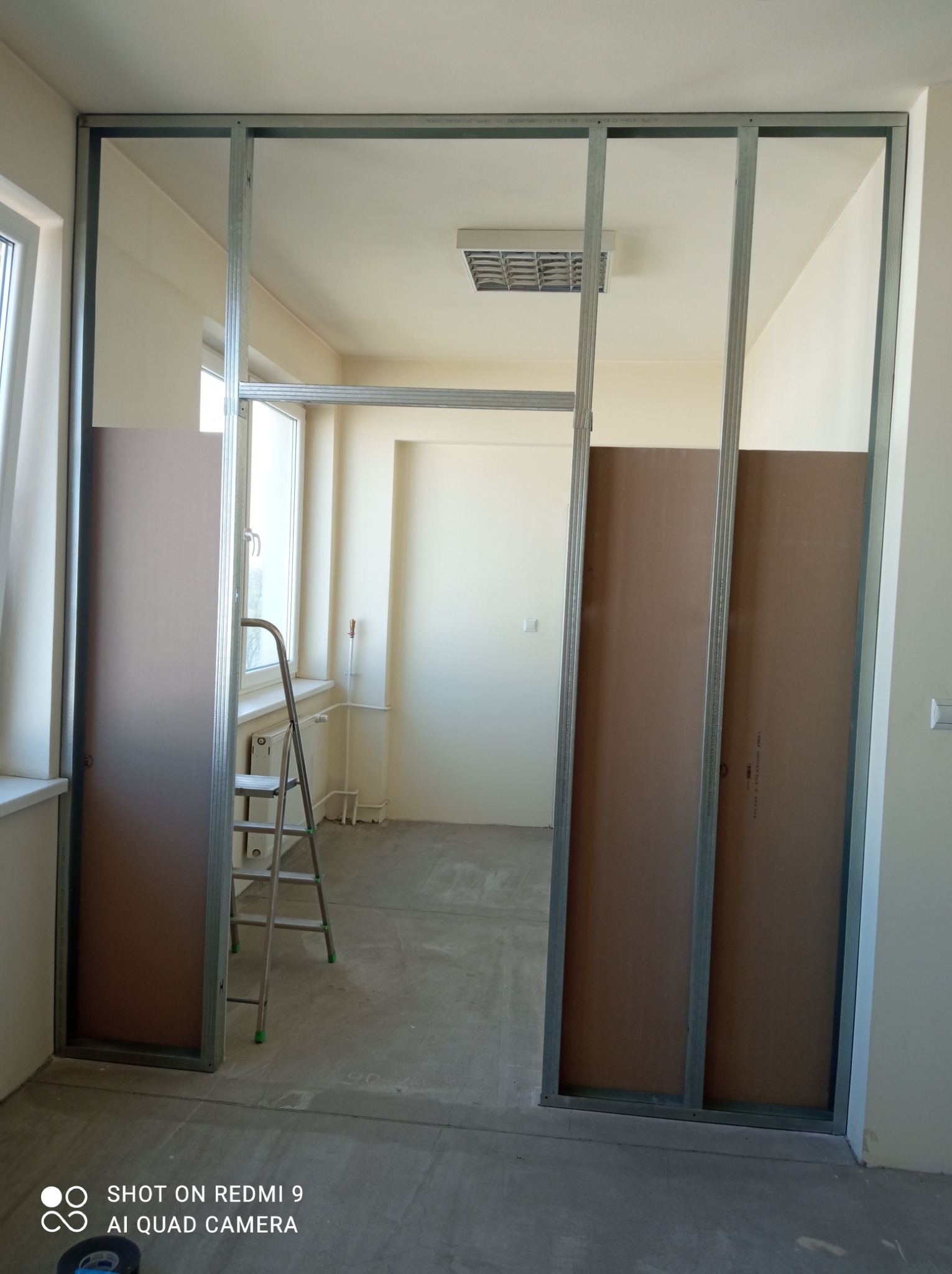 Montaż ścianki działowej z płyt kartonowo-gipsowych na metalowym stelażu, widoczna drabina aluminiowa i fragment okna.