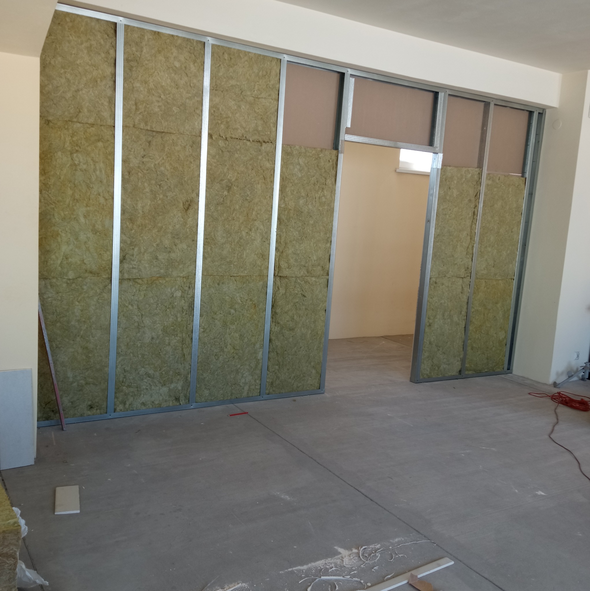 Konstrukcja ścianki działowej z metalowych profili, częściowo wypełniona wełną mineralną, widoczny otwór drzwiowy i przygotowane płyty kartonowo-gipsowe. Podłoga betonowa, surowa, z widocznymi...