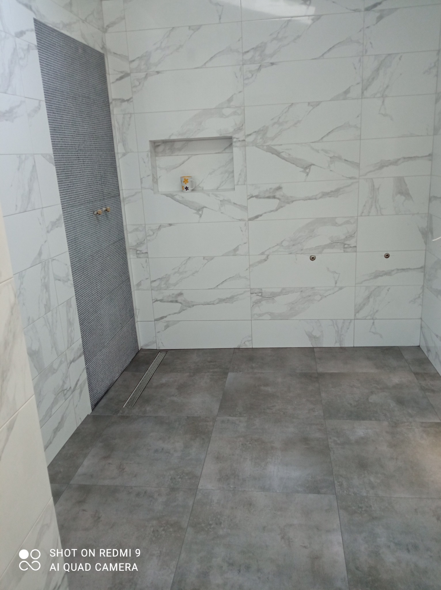 Wykończona łazienka z szarymi płytkami podłogowymi i białymi marmurkowymi płytkami ściennymi, wnęka z dekoracją i odpływ liniowy.