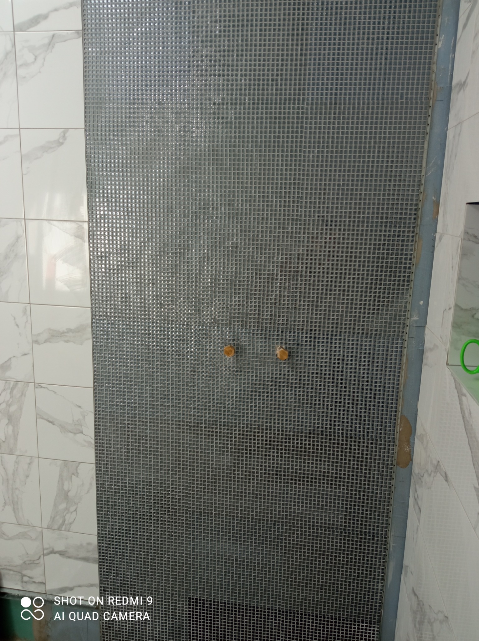 Ściana wyłożona mozaiką z małych, srebrnych płytek kwadratowych, z dwoma drewnianymi gałkami w centralnej części, obok ściany z białych płytek imitujących marmur. Widoczne ślady kleju montażowego...