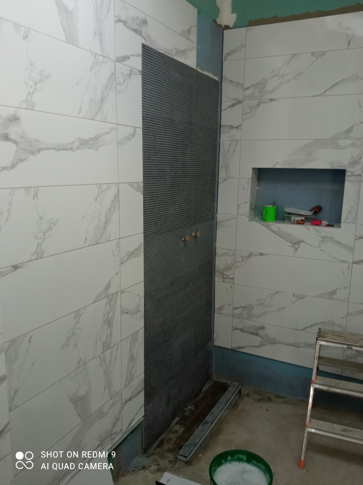 Ściana łazienki w trakcie wykańczania z białymi płytkami imitującymi marmur, widoczna siatka pod płytki w strefie prysznica, aluminiowa drabina i wiadro z zaprawą.