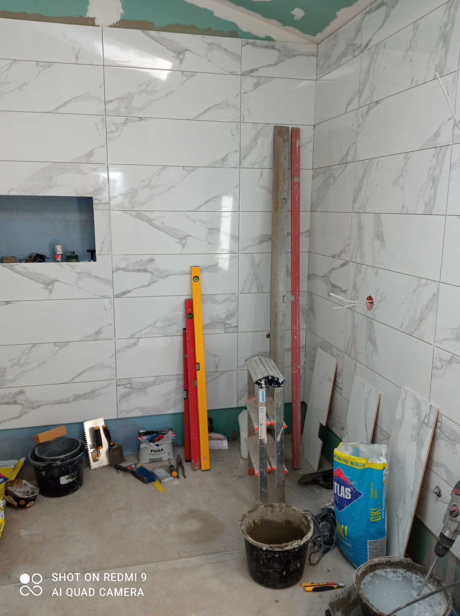 Ściany łazienki wyłożone białymi płytkami imitującymi marmur, z widocznymi narzędziami glazurniczymi, poziomnicami, wiadrami z zaprawą i workiem kleju Atlas, w trakcie prac wykończeniowych.