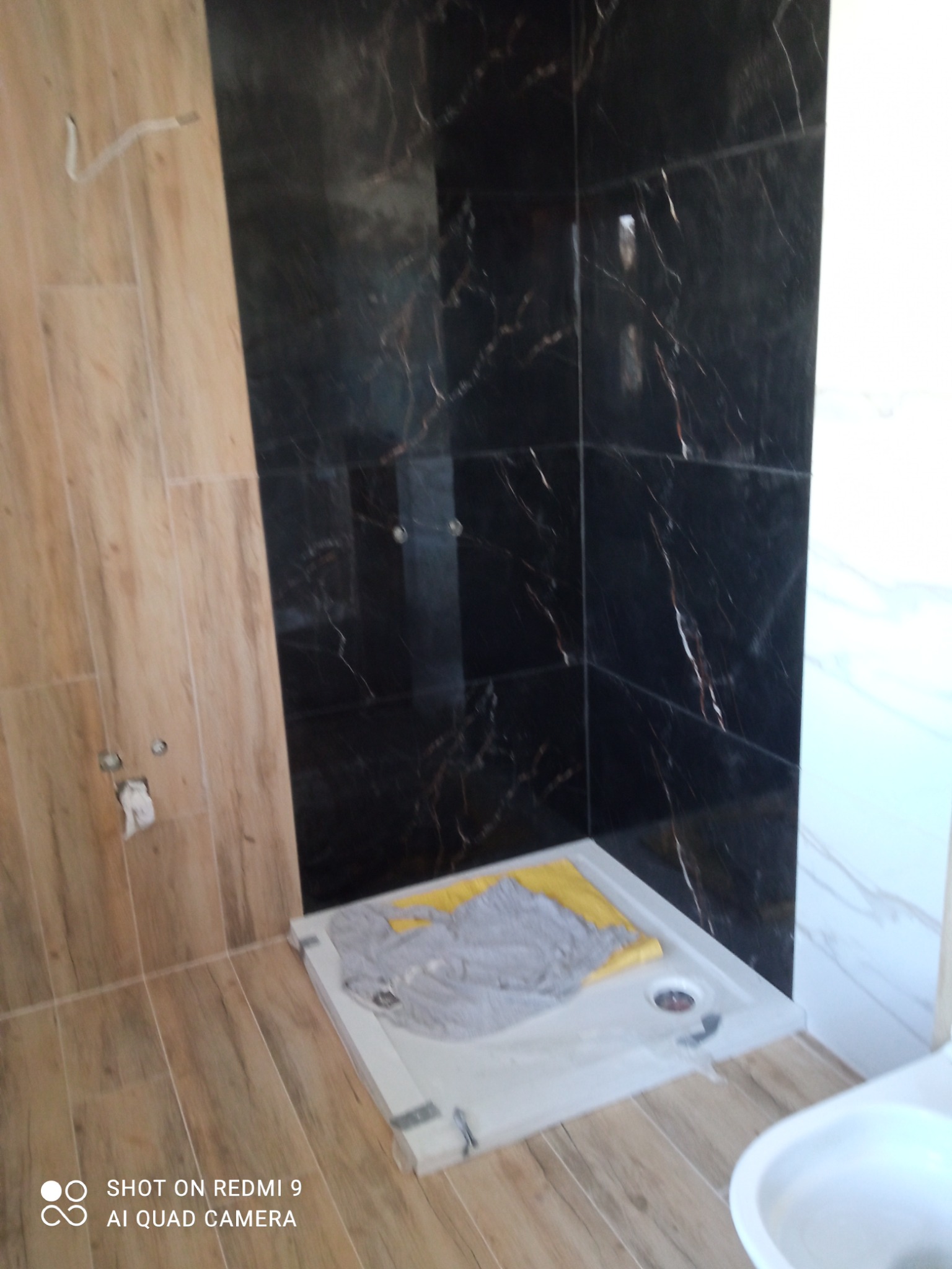 Niedokończony prysznic z czarnymi marmurkowymi płytkami i drewnopodobnymi płytkami podłogowymi, zabezpieczony szarą i żółtą szmatą, widoczny fragment muszli klozetowej.