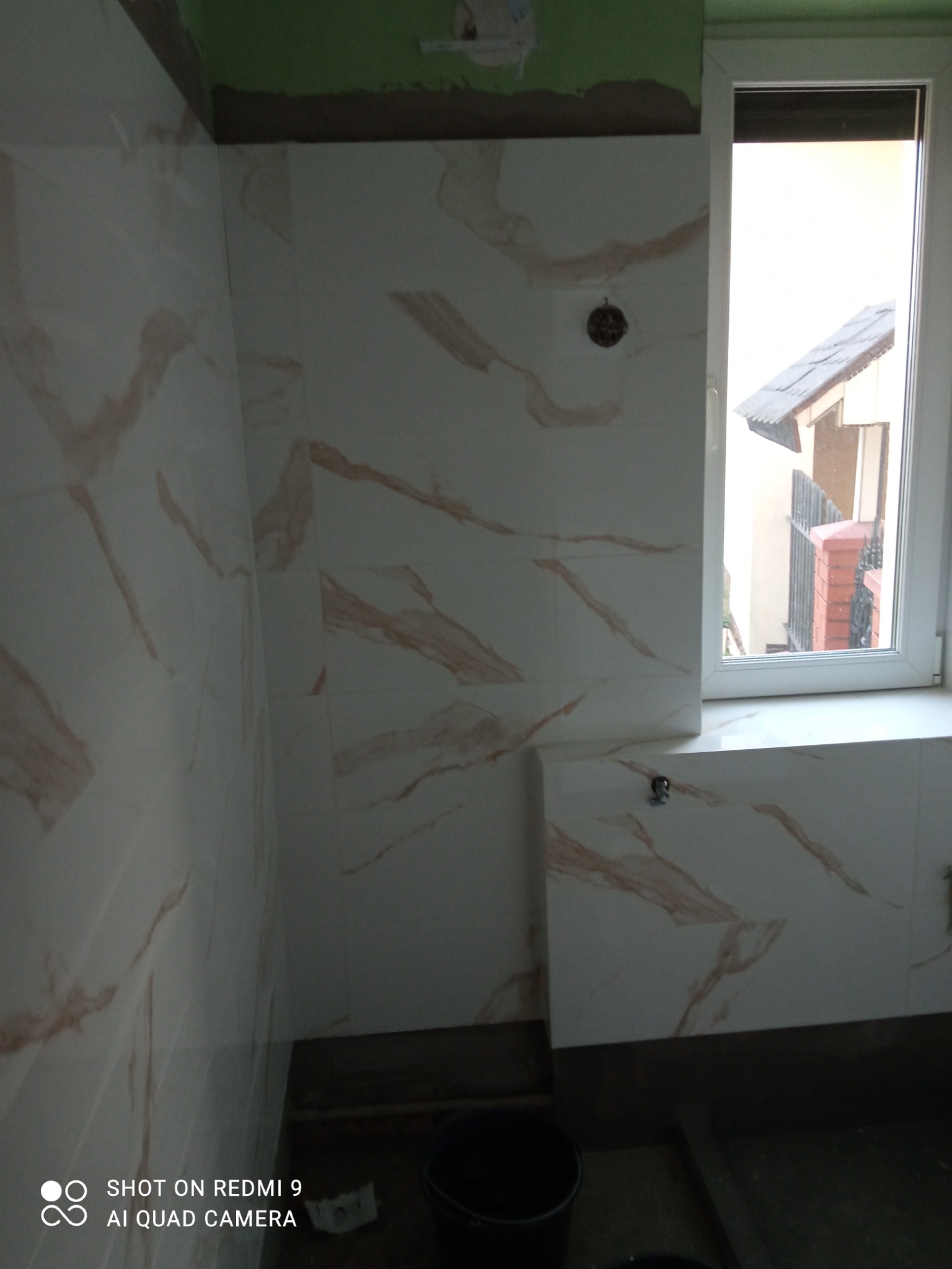 Ściany łazienki wyłożone białymi płytkami z marmurkowym wzorem w odcieniach beżu, widoczne niedokończone wykończenia przy suficie i oknie, czarne wiadro w rogu pomieszczenia.