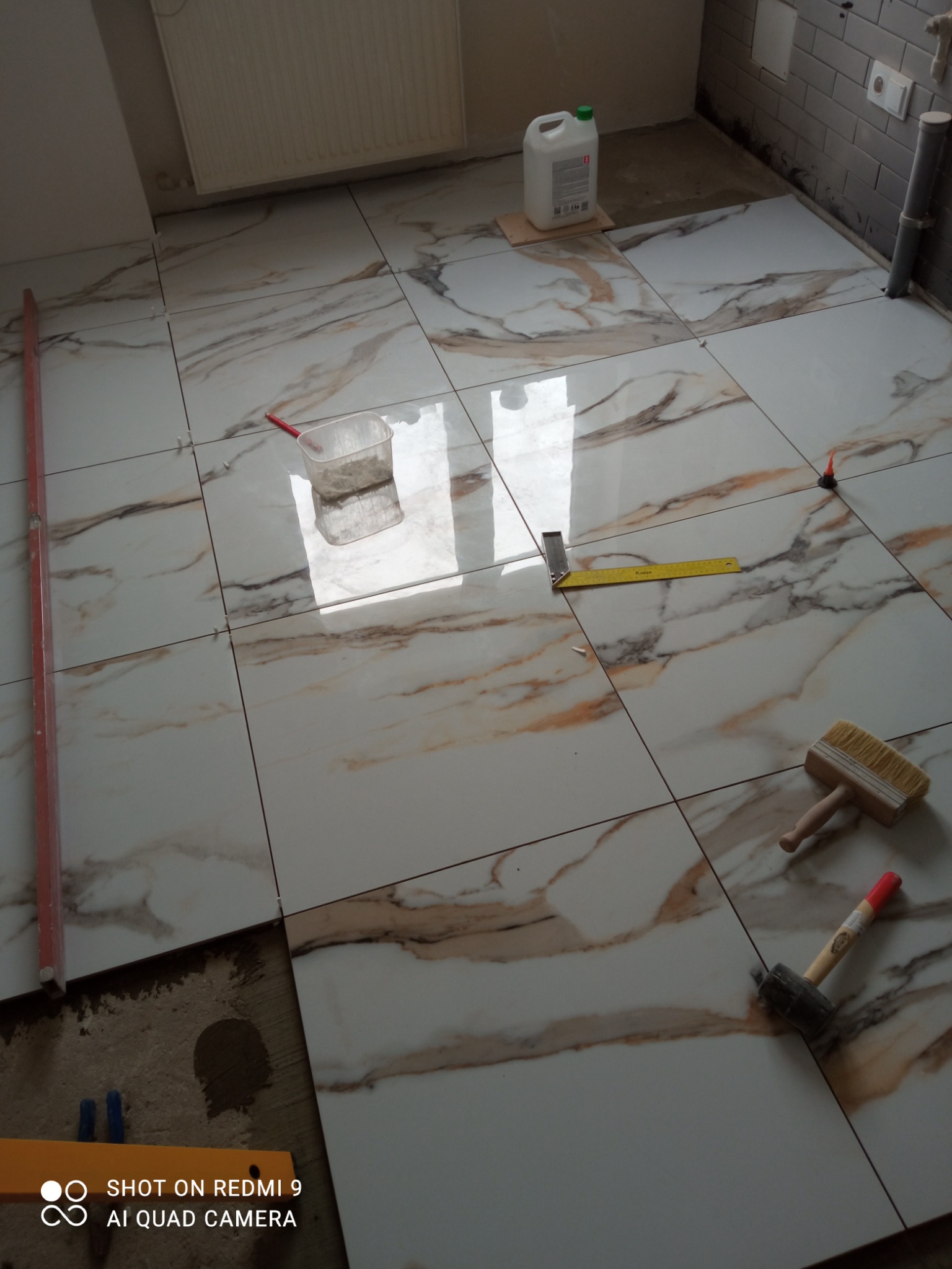 Układanie płytek podłogowych imitujących marmur, widoczne narzędzia glazurnicze: poziomica, kątownik, pędzel, gumowy młotek, pojemnik z zaprawą, butelka z płynem. W tle szare płytki ścienne.