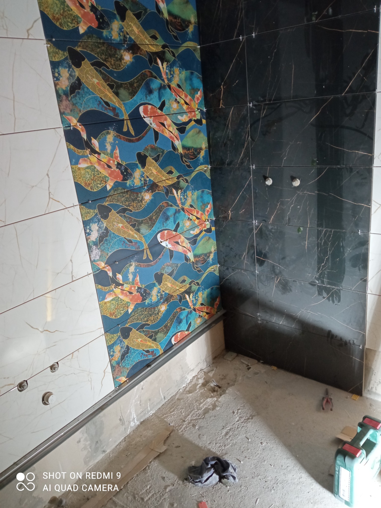 Ściana łazienki z częściowo ułożonymi płytkami: białe marmurkowe, niebieskie z motywem ryb koi i czarne z żyłkowaniem, widoczne narzędzia glazurnicze i niedokończona podłoga.