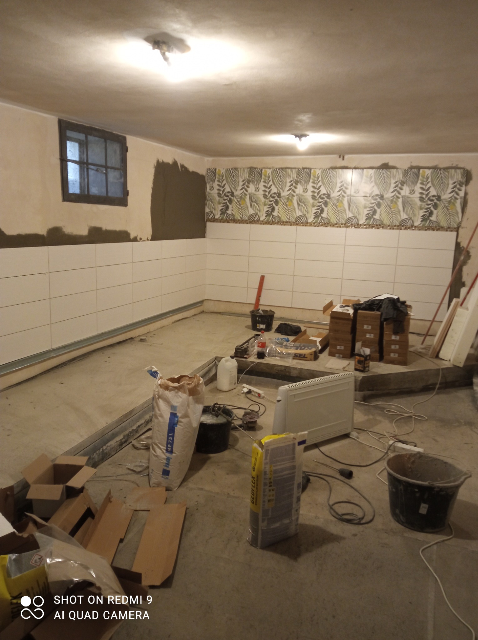 Wnętrze w trakcie układania białych płytek na ścianach, z fragmentem dekoracyjnego pasa płytek z motywem liściastym, widoczne worki z zaprawą, grzejnik elektryczny i narzędzia glazurnicze, okno...