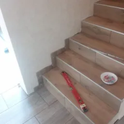 Wykończenie schodów płytkami imitującymi drewno z metalowymi listwami w Sędziszowie Małopolskim, widoczny poziomica i miseczka z krzyżykami dystansowymi.