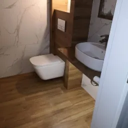 Nowoczesna łazienka z podwieszaną toaletą, umywalką nablatową i panelami imitującymi drewno na podłodze oraz ścianie, wykończona marmurowymi płytkami.