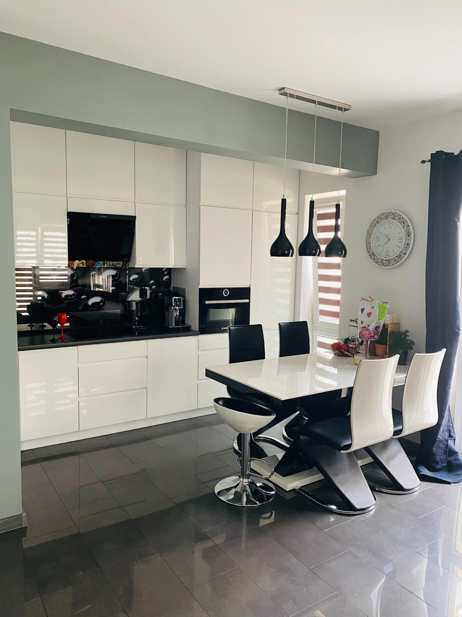 Nowoczesna kuchnia z białymi szafkami, czarnym blatem i błyszczącą podłogą z szarych płytek; stół z białym blatem i czarno-białymi krzesłami.