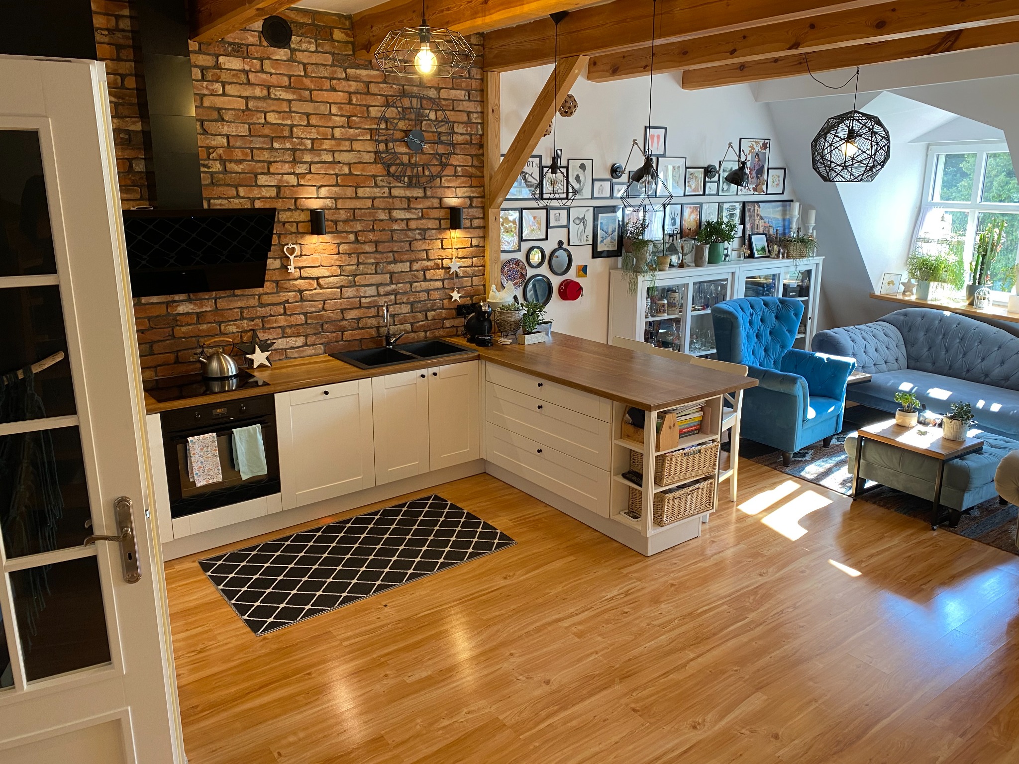 Jasna kuchnia z ceglaną ścianą, drewnianymi belkami stropowymi i narożną zabudową, połączona z salonem w stylu eklektycznym z błękitnymi fotelami i sofą.