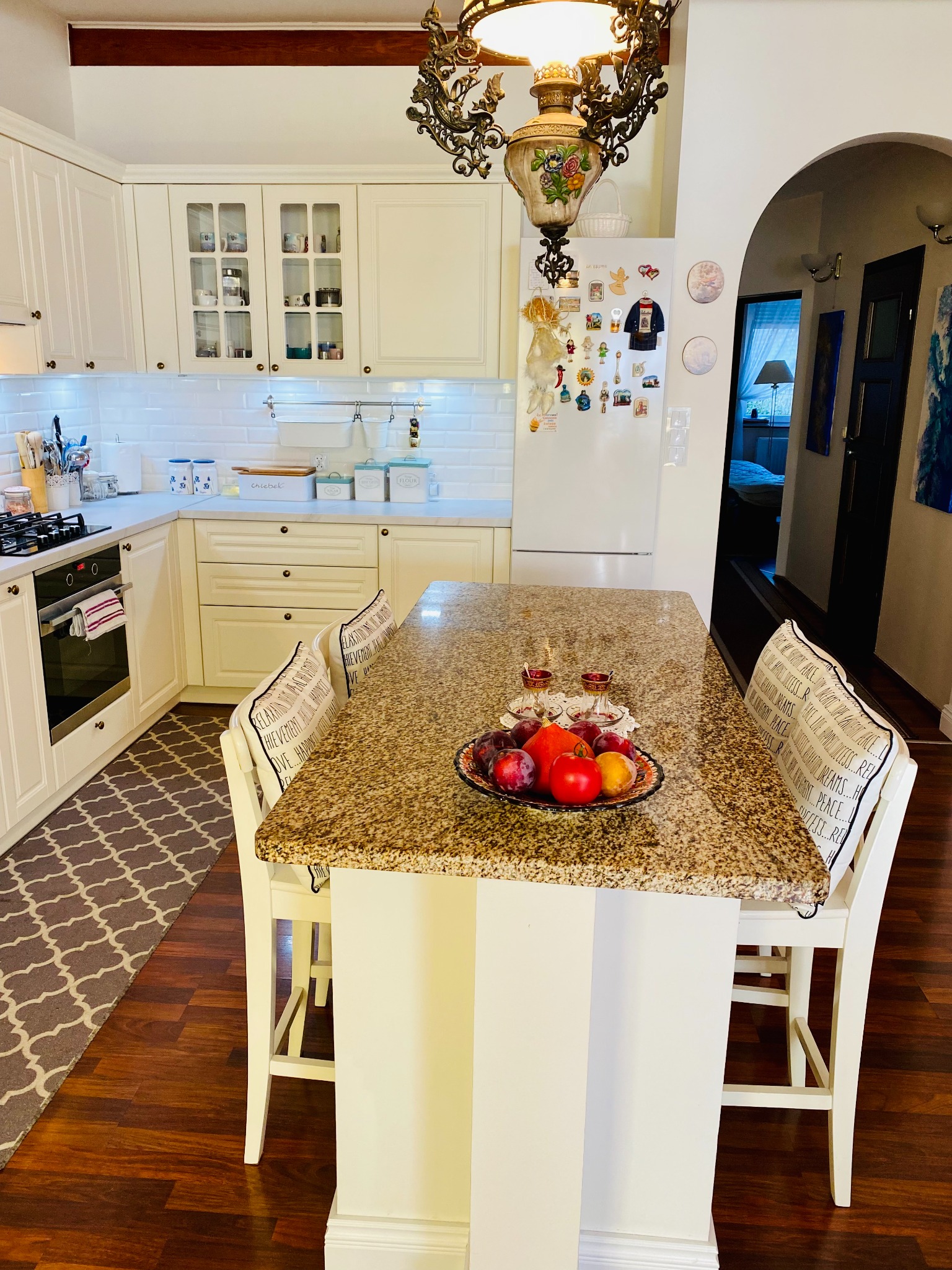 Jasna kuchnia z wyspą wykończoną blatem z granitu, na której leżą owoce w ozdobnej misie i dwie szklanki do herbaty, w tle białe szafki i lodówka z magnesami.