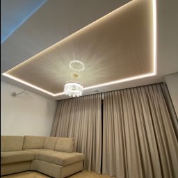 Wykończenia z klimatem - Salon z beżową, narożną sofą i podwieszanym sufitem z oświetleniem LED oraz kryształowym żyrandolem.