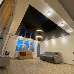 Wykończenia z klimatem - Salon z czarnym, błyszczącym sufitem napinanym, oświetleniem punktowym i nowoczesną lampą wiszącą, widoczne okno z widokiem na zewnątrz i podłoga z paneli.