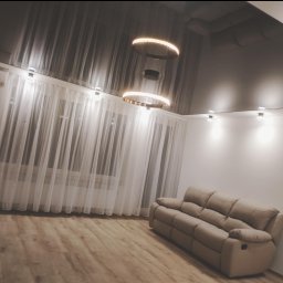 Wykończenia z klimatem - Salon z ciemnym, błyszczącym sufitem napinanym, okrągłymi lampami LED, jasnymi zasłonami i beżową sofą z funkcją relaks.