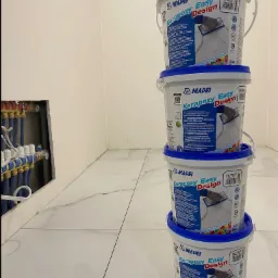 Stos czterech wiader z epoksydową zaprawą do spoinowania płytek, marki Mapei Kerapoxy Easy Design, na tle wykończonej białymi płytkami podłogi i fragmentu instalacji hydraulicznej w ścianie.