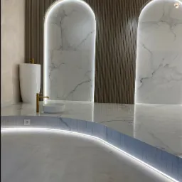 Nowoczesna łazienka z podświetlanym podestem, marmurowymi płytami w kształcie łuków i wolnostojącą umywalką w kolorze białym z złotą armaturą.