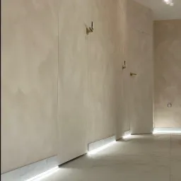 Beżowe ściany z dekoracyjnym tynkiem i ukrytymi drzwiami z złotymi klamkami, podświetlenie LED przy podłodze, marmurowe płytki podłogowe.