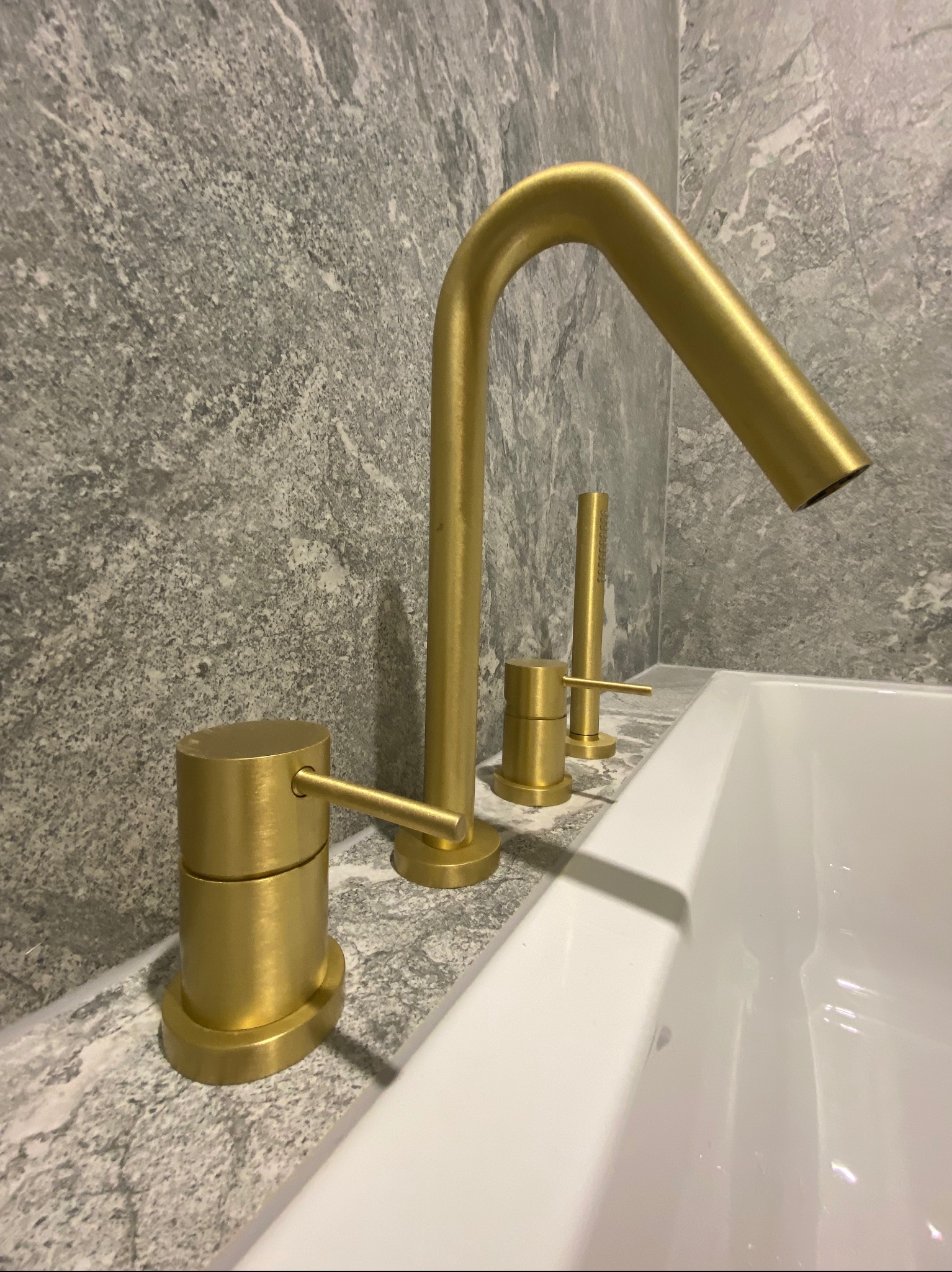 Złota armatura wannowa z baterią i słuchawką prysznicową na marmurowym blacie w łazience wykończonej szarą płytką imitującą kamień.