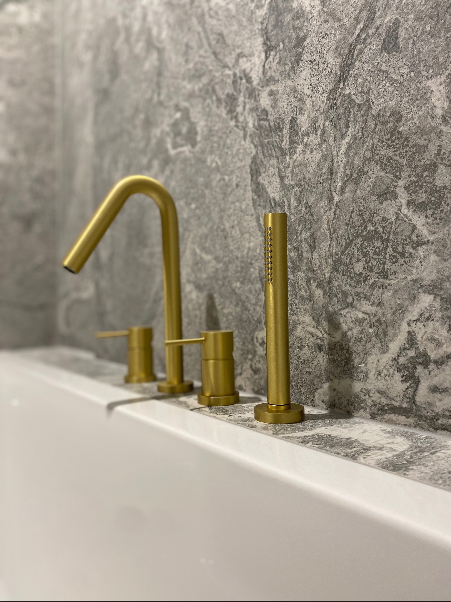 Bateria wannowa w kolorze szczotkowanego złota z deszczownicą na tle szarej, marmurkowej ściany.