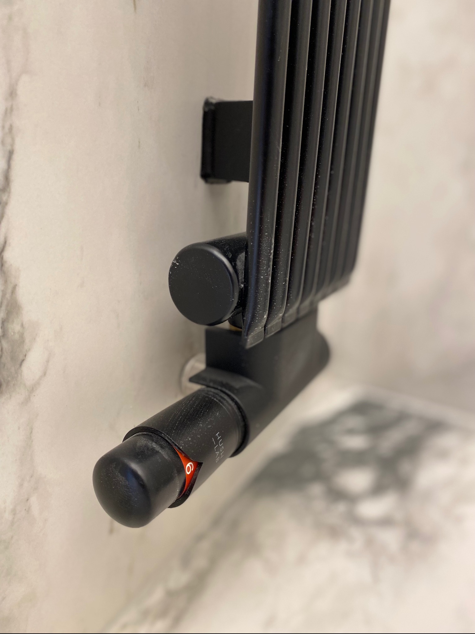 Czarny grzejnik łazienkowy drabinkowy z termostatem ustawionym na cyfrę 6, zamontowany na ścianie wykończonej płytkami imitującymi marmur.